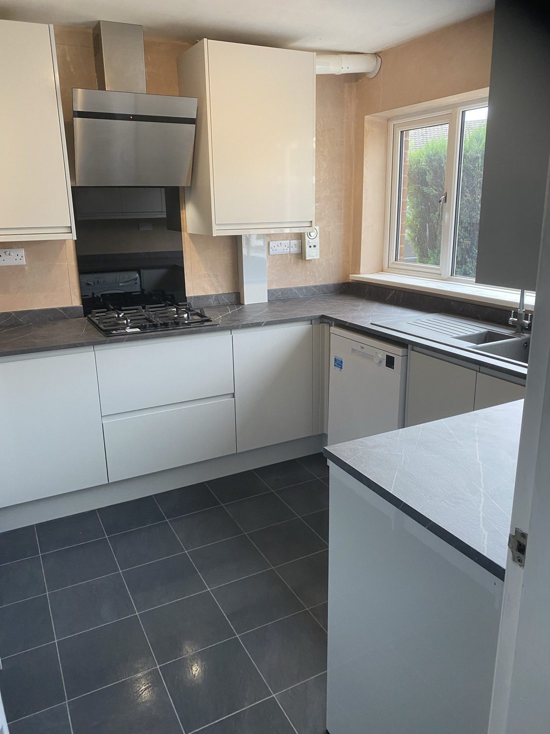 A soft cream handleless kitchen with a marbled worktop, stainless extractor and gas hob, laid over a dark slate-tone floor — clean, bright and easy to live with.